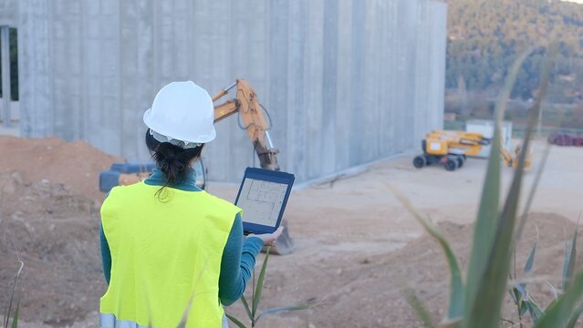 Woman architect or engineer wearing a hard hat and safety vest, reading a building blueprint on a tablet, overseeing construction site development and progress