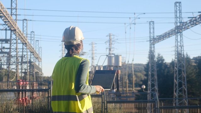 Engineer wearing safety vest and hard hat, holding a laptop, standing with her back to the camera at an outdoor electrical power substation monitoring the grid