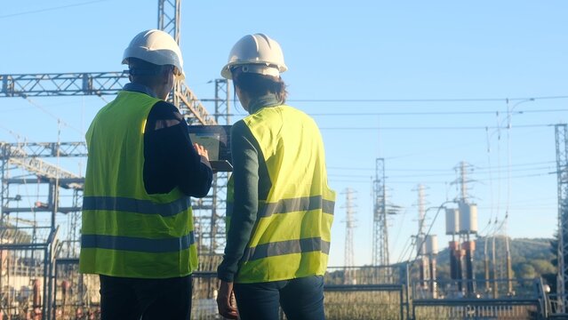 Engineers collaborating and checking system parameters on a laptop, standing in front of a high-voltage electrical substation, ensuring power grid stability and energy distribution reliability