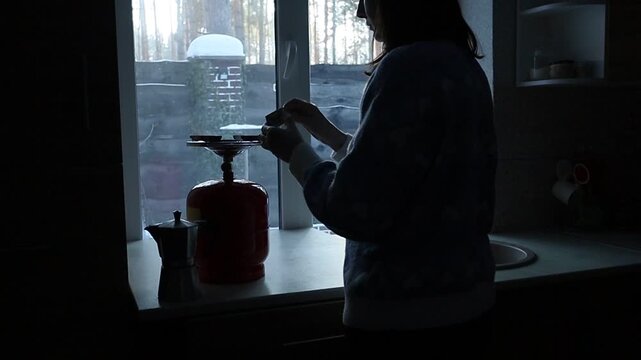 A young Ukrainian woman lights a gas stove with a match to make herself coffee during a power outage. The intimate kitchen scene highlights everyday resilience and adaptation to electricity shortages 