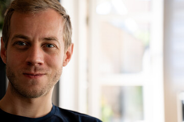 Naklejka premium A blond haired man with blue eyes and light stubble smiles gently, looking forward. He is situated indoors, illuminated by natural daylight from a nearby window
