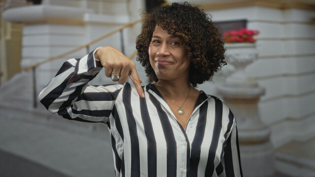 Woman in striped shirt wearing gold pendant smiling with finger pointing to head on street; idea happiness.
