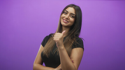 Obraz premium Young hispanic woman showing thumbs up with hand and crossed arms in studio with purple backdrop; confidence approval.