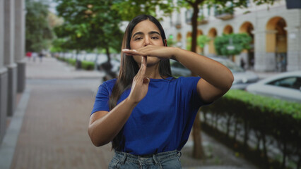 Fototapeta premium Young hispanic woman makes timeout hand sign near face on a city street lined with trees and arched building facades; pause assertive boundary.