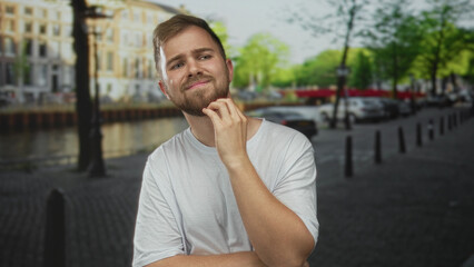 Fototapeta premium Man in white t shirt touching chin and scratching beard on amsterdam street beside a canal and parked cars; pensive doubt.