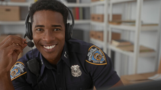 Man police officer smiles and adjusts headset microphone while speaking at a desk in a building with shelves and boxes, badge visible; professional duty readiness.