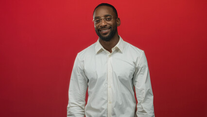 Obraz premium Man adjusting round eyeglasses with hands near face wearing white shirt in studio against red backdrop; confidence.