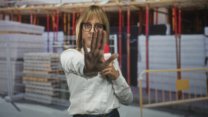 Fototapeta premium Woman reporter holds microphone and shows palm forward stop gesture in building site facing camera and raising hand to block access; defiance safety warning.