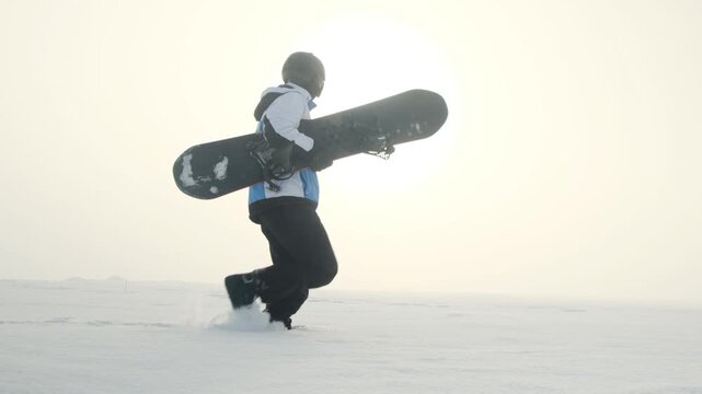 Camera follows from side a snowboarder walking on snow carrying snowboard in hands. Fully equipped rider ready for descent from mountain peak. Snowboarding as winter recreation and extreme sport