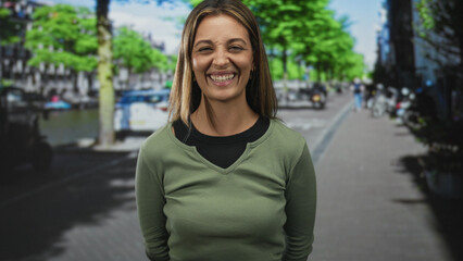 Fototapeta premium Woman smiling with visible face and teeth, wearing green sweater and gold earrings, standing on an amsterdam street by a canal with sunlit trees, hands behind back; joy urban connection.