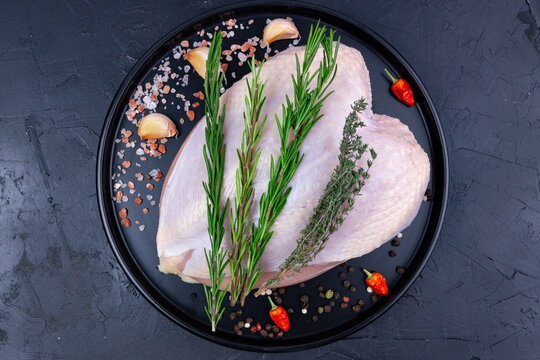 Raw chicken breast is placed on a black plate with herbs and spices in a kitchen setting during the daytime