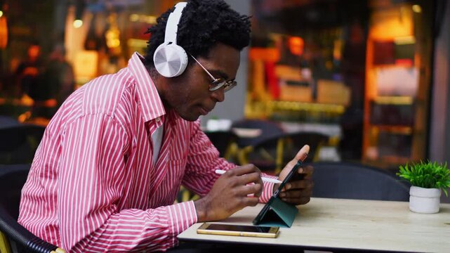 Side view of young Black man using graphic tablet with pen while listening to music in white headphones, creative professional working remotely in urban environment, digital art concept
