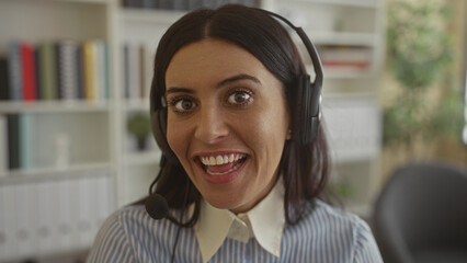 Woman wearing headset and microphone speaking and smiling, visible teeth, striped blouse, seated at desk inside office building with bookshelves and plant  warmth connection. © Krakenimages.com