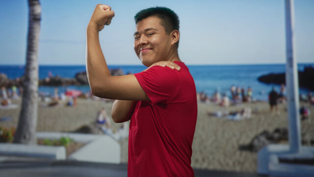 Man flexes arm and holds shoulder on street promenade by the beach while smiling with closed eyes; confidence summer fun.