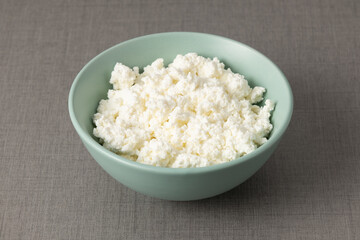 Fresh cottage cheese sits in a light blue bowl on a gray tablecloth during midday sunlight