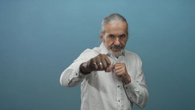 Man with hoary long hair and beard throwing punches with fists in studio blue backdrop wearing white shirt; determination grit.