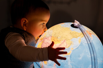 A curious little boy, a few months old, looks at a glowing globe lamp in a dark room and touches the map with his hand, exploring the geography of the Earth, future generations.
