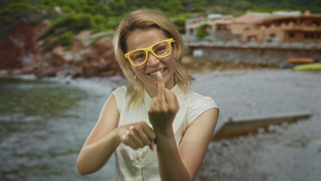 Woman wearing yellow glasses shows middlefinger at sunlit building by seaside; raw rebellious defiance.