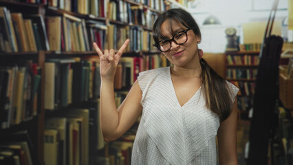 Woman with glasses and ponytail showing rock horns hand gesture in a library building with bookshelves and stacked books  playful curiosity. © Krakenimages.com