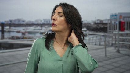 Obraz premium Woman touches her neck, pushes dark hair behind her ear, and closes her eyes on a port street by fishing boats; serenity.