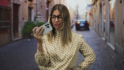 Obraz premium Woman holding smartphone at arm's length, frowning and squinting while checking the screen in a narrow cobblestone street lined with buildings and parked cars; annoyance.