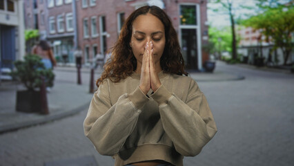 Fototapeta premium Woman with hands pressed together in prayer on a cobblestone street, cropped sweatshirt visible; serenity reflection.