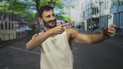 Fototapeta premium Man points finger to smartphone on street beside canal boat while taking a selfie and smiling broadly; casual confidence.
