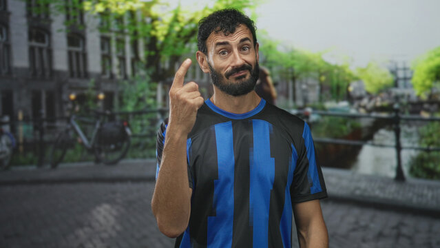 Man in soccer jersey points finger with a slight smirk standing on a street canal bridge beside bicycles and cobblestone; cheeky confidence.