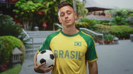Young man wearing a brasil soccer jersey holding a soccer ball and rubbing his eye on a street with landscaped steps and planters; disappointment.
