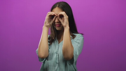 Naklejka premium Woman framing her eyes with hands to make binoculars in studio with purple backdrop; curiosity exploration.