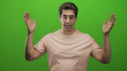 Naklejka premium Man raising both hands with palms open and smiling, wearing glasses and a light pink t shirt against a studio green screen, expressive pose; joy.