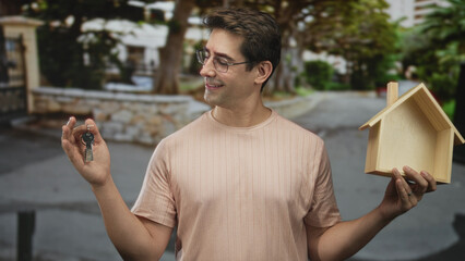 Fototapeta premium Man holds keys in left hand and a wooden house model in right hand on a street, smiling and wearing glasses; homeownership pride.