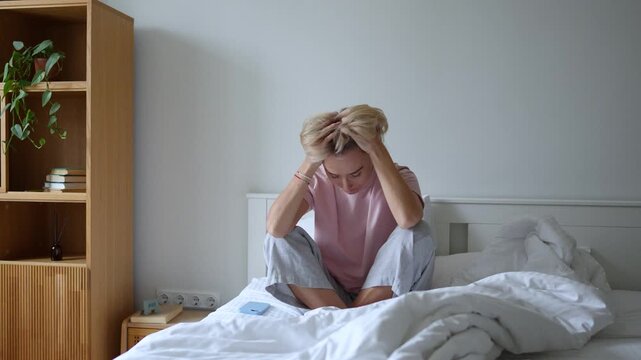 Middle aged woman with exhausted expression woke up after sleepless night or bad dream, sitting in bed in bedroom feeling morning blues, checking time and messages on smartphone, tiredness and fatigue