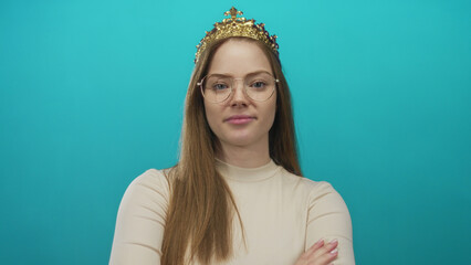 Woman wearing a gold crown and glasses stands with arms crossed in studio against a turquoise wall  confidence. © Krakenimages.com