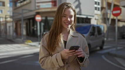 Obraz premium Young caucasian woman smiling while tapping smartphone with both hands on a city street wearing a trench coat; connection focus engagement.