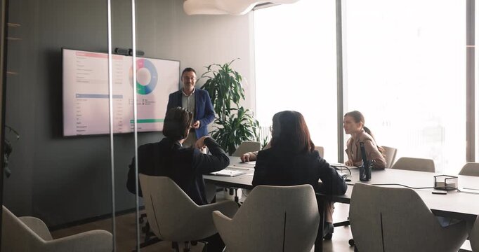 Middle-age leader make presentation stands next to screen displaying financial graphs, discuss budget allocation, performance charts during corporate meeting with team. Management, seminar in office