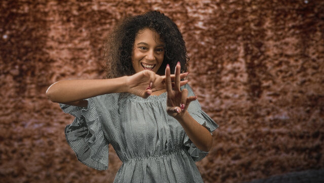 Woman forming a hashtag sign with her hands while smiling, wearing a gingham dress and curly hair against a textured backdrop in a studio setting; joy self expression.