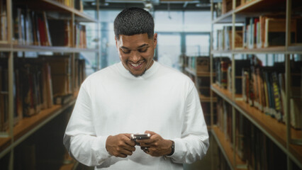Man with beard in white shirt holding smartphone with both hands and smiling while typing between bookshelves in library building  relaxed contentment. © Krakenimages.com