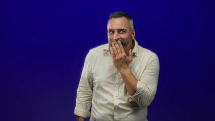 Middle aged hispanic man covering mouth with hand in studio against deep purple backdrop; surprise candid connection.