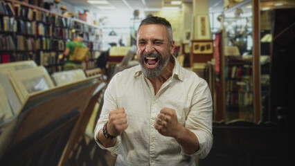 Man with clenched fists shouting in a library among bookshelves and records, grey hair and beard, fists raised in celebration  victory joy relief. © Krakenimages.com