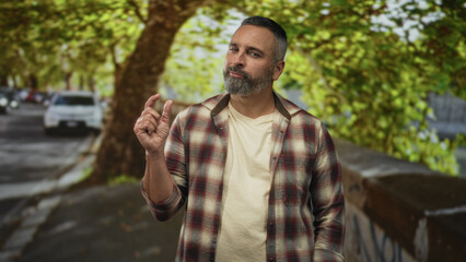 Man displaying a pinch gesture with one hand on a tree lined street near parked cars, wearing a plaid shirt and beard; quiet skepticism.