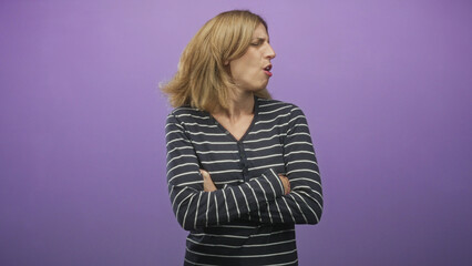 Fototapeta premium Woman with mouth puckered and arms crossed, head turned and hair in motion, wearing striped top against purple studio backdrop; dismissal annoyance.