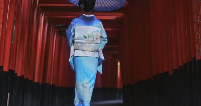 Cinematic slow motion as Japanese woman in kimono walks down long line of old torii gates with umbrella
