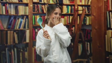 Young woman chef in white coat holds turquoise ladle and covers mouth with hand while smiling amid bookshelves in a library building  playful joy. © Krakenimages.com