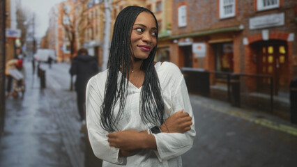 Fototapeta premium Black woman smiles with long braids and white blouse, pendant and watch visible while posing on a wet city street; confidence candid urban.