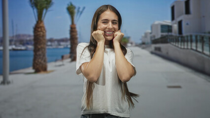 Fototapeta premium Teenage girl showing braces and raised fists on a sunny street by palm trees and marina, smiling broadly while walking toward camera; youthful joy.