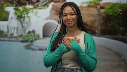 Fototapeta premium Woman standing with hands over chest near a hotel pool and white arch building, eyes closed, braided hair visible, green cardigan and casual jeans; gratitude.