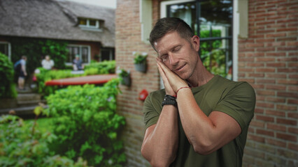 Fototapeta premium Man resting head on hands with hands pressed to cheek at a house building entrance, sleep gesture; calm rest.