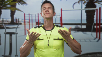 Man with palms up gesture and whistle necklace in gymnasium by outdoor calisthenics bars; calm...