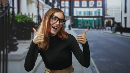 Fototapeta premium Woman with glasses showing thumbs up and exposed midriff on a street lined with buildings and railings; confidence success.